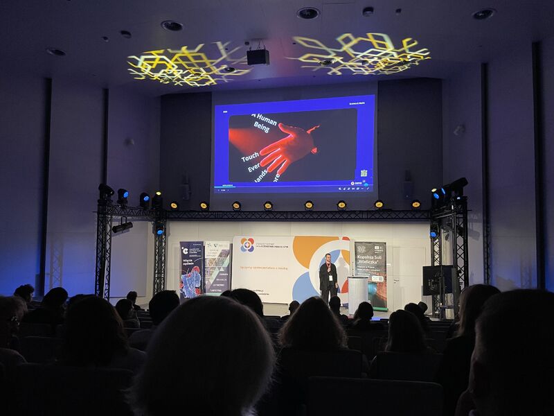 Kamil Pilch of Sano speaking on stage at SPIN 2026, with a glowing red hand on the screen and the audience in front