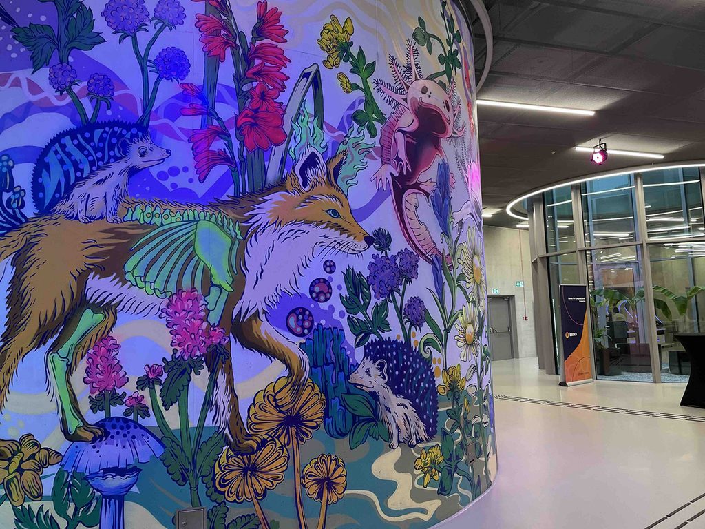 Colourful mural of a fox, hedgehogs and an axolotl among flowers in the Cogiteon foyer, with a Sano roll-up nearby